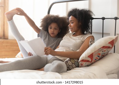 African American Family Mom And Cute Little Kid Daughter Read Book Fairy Tale Sitting In Bed, Small Preschool Child Girl Listening To Black Mom Learning Reading Relaxing In Bedroom In The Morning