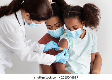 African American Family Mother And Cute Daughter Making Vaccination At Clinic, Wearing Protective Face Masks. Doctor Putting Band On Kid Shoulder After Injection Against Coronavirus, Closeup