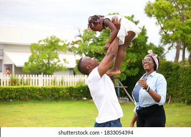 African American Family Playing With Young Happy Little Daughter On Green Grass Field While Enjoying Summer Garden Outside The House In The Neighborhood With Copy Space.