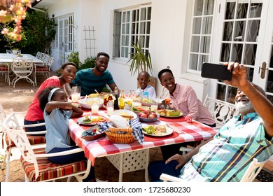 African American family spending time together in the garden on a sunny day, sitting by a table and having a lunch, while a senior man is taking a selfie with his smartphone.  - Powered by Shutterstock