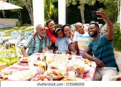 African American family spending time together in the garden on a sunny day, sitting by a table and having a lunch, while a man is taking a selfie with his smartphone.  - Powered by Shutterstock