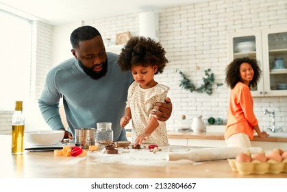 African American Family With Their Little Daughter With Curly Fluffy Hair Having Fun And Cooking Pastries In The Kitchen. Dad And Daughter Cooking Together And Cutting Out Cookies From The Dough.