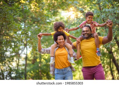 African American family walking trough park. Parents carrying children on piggyback. 