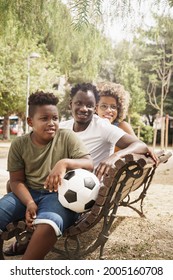 African American Family With Young Cool Millennial Parents And Cute Smiling Kid Holding A Soccer Ball Sitting On Bench In Outdoor Public Park. Focus On Center Black Man