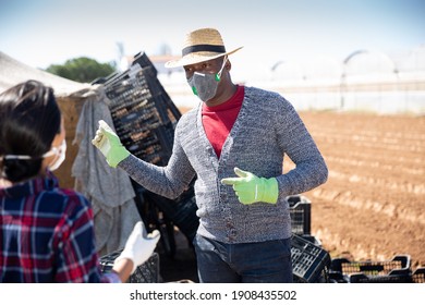 African American Farmer Wearing Protective Face Mask Talking To Hispanic Female Worker On Farm Field On Sunny Spring Day. Concept Of Precautions And Social Distancing In Coronavirus Pandemic