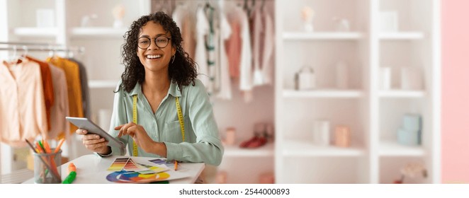 African American fashion designer smiles as she reviews designs on a tablet in a well-organized studio. Colorful fabrics hang nearby, showcasing her artistic environment. - Powered by Shutterstock