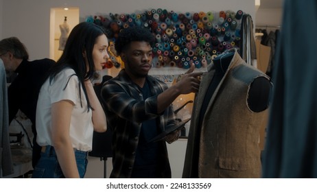 African American fashion designer talks with seamstress in atelier and shows sketch of future suit on digital tablet computer. Mannequin in unfinished tailored suit. Concept of fashion and hand craft. - Powered by Shutterstock