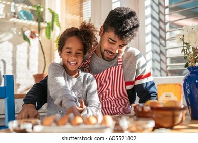 African American Father And Daughter Cooking In The Kitchen. Dad Is Wearing An Apron. They Are Both Smiling.