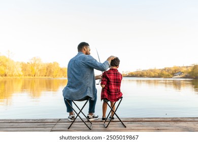 An African American father and his son sit on folding chairs on a wooden pier near a lake. The father, in a denim jacket and jeans, strokes his son's head while holding a fishing rod. The son, in a - Powered by Shutterstock