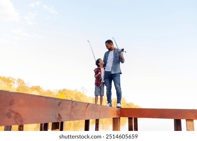 An African American father and his son stand on a wooden pier by a lake. The father, wearing a denim jacket and jeans, holds a fishing rod and hugs his son, looking at him with a smile. The son - Powered by Shutterstock