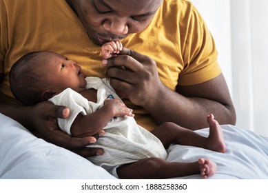 An African American father kissing hand,  his 12-day-old baby newborn son lying in bed in a white bedroom, with happy, concept to African American family and newborn