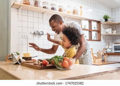 African American Father and little son making video call with digital tablet and waving hands laughing talking with family while cooking in kitchen. Black family have fun while pandemic virus at home