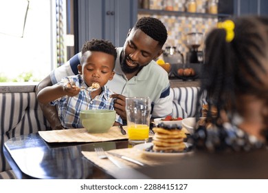 African american father looking at son eating breakfast cereals at dining table at home, copy space. Unaltered, family, togetherness, childhood, lifestyle, food, fresh and healthy concept. - Powered by Shutterstock