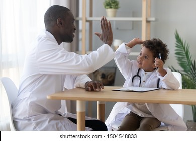 African American Father Playing Doctor With Little Adorable Son, Giving High Five, Cute Child Wearing White Uniform And Stethoscope Holding Pen, Sitting At Desk With Clipboard, Having Fun With Dad