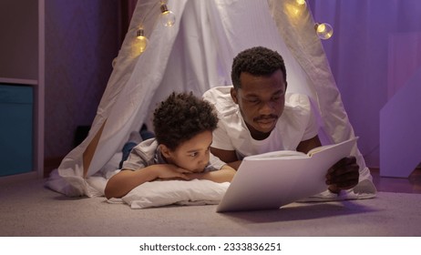African American father reads fairytale to delighted preschooler son in wigwam.