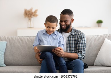 African American Father And Son Using Tablet Computer Browsing Internet Together Sitting On Couch At Home. Weekend Family Leisure, Gadgets And Modern Lifestyle Concept
