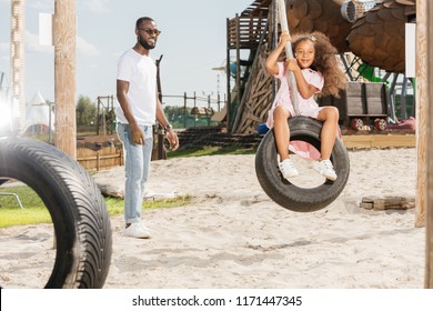 African American Father Standing Near Smiling Daughter On Tire Swing At Amusement Park