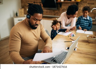 African American Father Working On A Computer And Going Through Reports While His Wife Is Homeschooling Their Kids. 