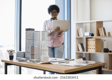 African American female architect develops project for modern city using digital laptop. Focus on building model on table. Concept of innovation and urban design.