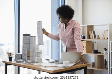 African American female architect in office setting holding building model. Young professional develops modern city project, focusing on urban planning and architectural design.
