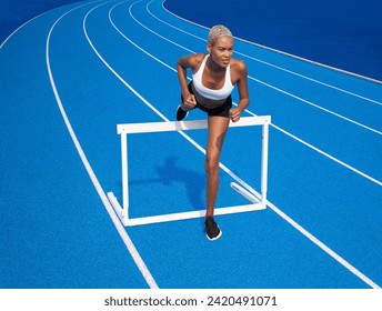 An African American female athlete is running on the track executing the hurdle jump over the blue lanes concept of race training and dedication in sports - Powered by Shutterstock