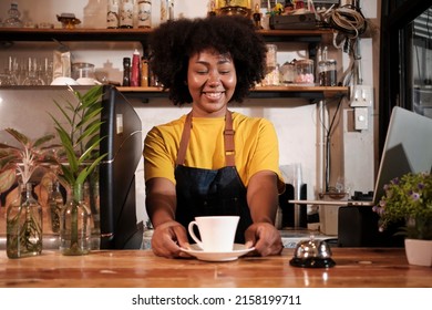 African American Female Barista Offers Cup Of Coffee To Customer With Cheerful Smile, Happy Service Works In Casual Restaurant Cafe, Young Small Business Startup Entrepreneur.