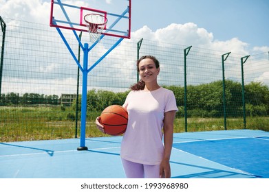 African American Female Basket Ball Player Standing On A Playground And Smiles Holding A Basketball Under Arms