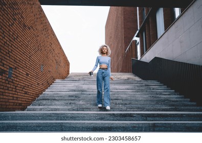 African American female in casual clothes walking downstairs near brick wall with paper coffee cup in hands and looking at camera in city street on weekend