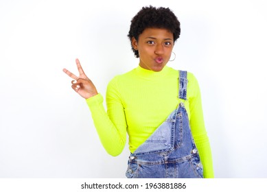 African American Female With Curly Bushy Wears Jeans Overalls Over White Wall Makes Peace Gesture Keeps Lips Folded Shows V Sign. Body Language Concept