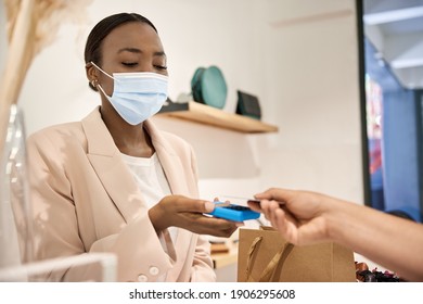 African American Female Customer In A Protective Face Mask Using Her Smart Phone To Make A Contactless Payment From In A Store