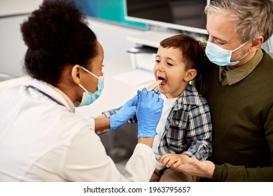 African American female dentist examining teeth of a small boy who is sitting on father's lap during dental appointment.  Focus is on boy.