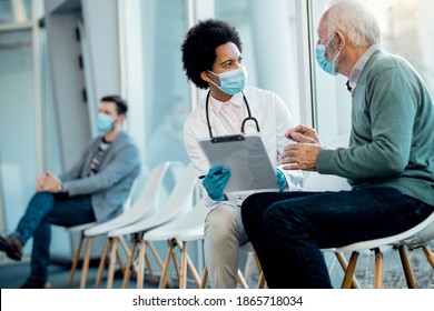 African American Female Doctor Analyzing Medical Record Of A Senior Patient While Talking To Him In Hospital Waiting Room. They Are Wearing Face Masks Due To Coronavirus Pandemic. 