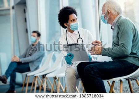 African American female doctor analyzing medical record of a senior patient while talking to him in hospital waiting room. They are wearing face masks due to coronavirus pandemic. 