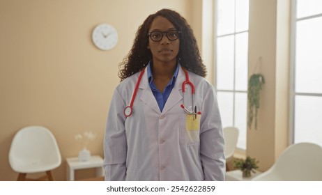 African american female doctor with curly hair and glasses standing in an indoor clinic lobby wearing a white coat and stethoscope - Powered by Shutterstock