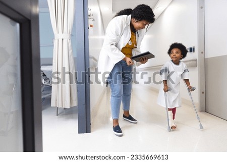 African american female doctor and girl patient walking with crutches in corridor at hospital. Hospital, medicine and healthcare, unaltered.