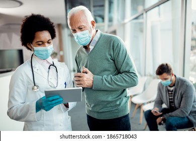 African American Female Doctor And Her Senior Patient Using Digital Tablet While Wearing Protective Face Mask At Hospital Hallway. 