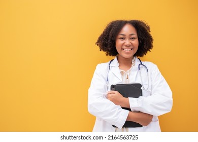 African American Female Doctor Holding Tablet Yellow Background. Smiling Mature Woman Doctor In White Lab Coat Standing Isolate. Successful Happy Healthcare Worker Looking At Camera, Copy Space.