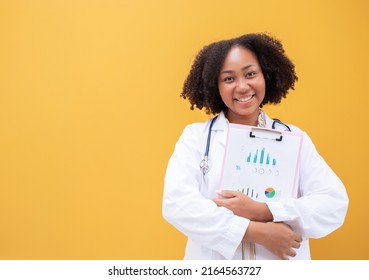 African American Female Doctor Holding Clipboard Yellow Background. Smiling Mature Woman Doctor In White Lab Coat Standing Isolate. Successful Happy Healthcare Worker Looking At Camera, Copy Space.