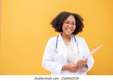 African American Female Doctor Holding Clipboard Yellow Background. Smiling Mature Woman Doctor In White Lab Coat Standing Isolate. Successful Happy Healthcare Worker Looking At Camera, Copy Space.