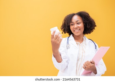 African American Female Doctor Holding Tablet Yellow Background. Smiling Mature Woman In White Lab Coat Standing Holding Medicine Pill Bottle Successful Happy Healthcare Looking At Camera, Copy Space.