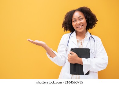 African American Female Doctor Holding Tablet Yellow Background. Smiling Mature Woman Doctor In White Lab Coat Standing Isolate. Successful Happy Healthcare Worker Looking At Camera, Copy Space.