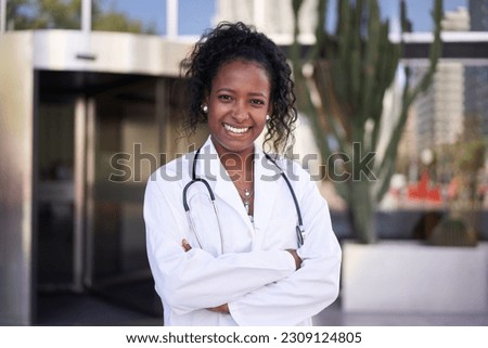 African American female doctor in lab coat with stethoscope outside clinic. Smiling young people medical specialty posing for photo outdoors . Happy and cheerful person with arms crossed.