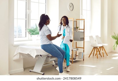 African American female doctor and patient talking at the clinic. Young woman sitting on a medical examination bed together with her friendly, smiling physician, asking questions and asking for advice - Powered by Shutterstock