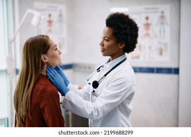 African American Female Doctor Touching Woman's Neck During Thyroid Exam At Medical Clinic.