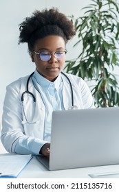 African American Female Doctor Using Laptop Computer For Medical Work Or Education. Young Physician Working At Desk In Office Writing Electronic Prescription. Online Chat With Patient, Telemedicine