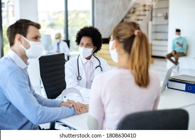 African American Female Doctor Wearing Protective Face Mask While Having Medical Appointment With A Couple At Doctor's Office. 