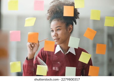 African American female employee write down on colorful sticky notes manage list, concentrated biracial woman work on startup brainstorm collaborate plan on stickers on glass wall - Powered by Shutterstock