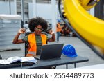 African american female engineer is stretching her arms while working on a laptop in a factory with a robotic arm in the background