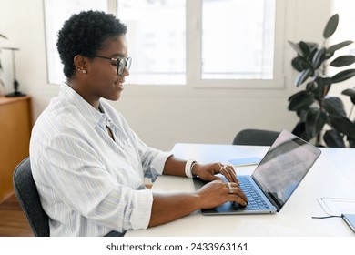 African American female freelancer with short hair sitting at the desk with laptop and typing, writing article, smiling and blogging, enjoying and creating design for new project, side view
