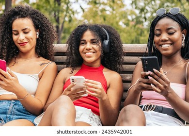 African american female friends sitting on a park bench laughing and having fun while using smartphone and listening to music with headphones - Powered by Shutterstock